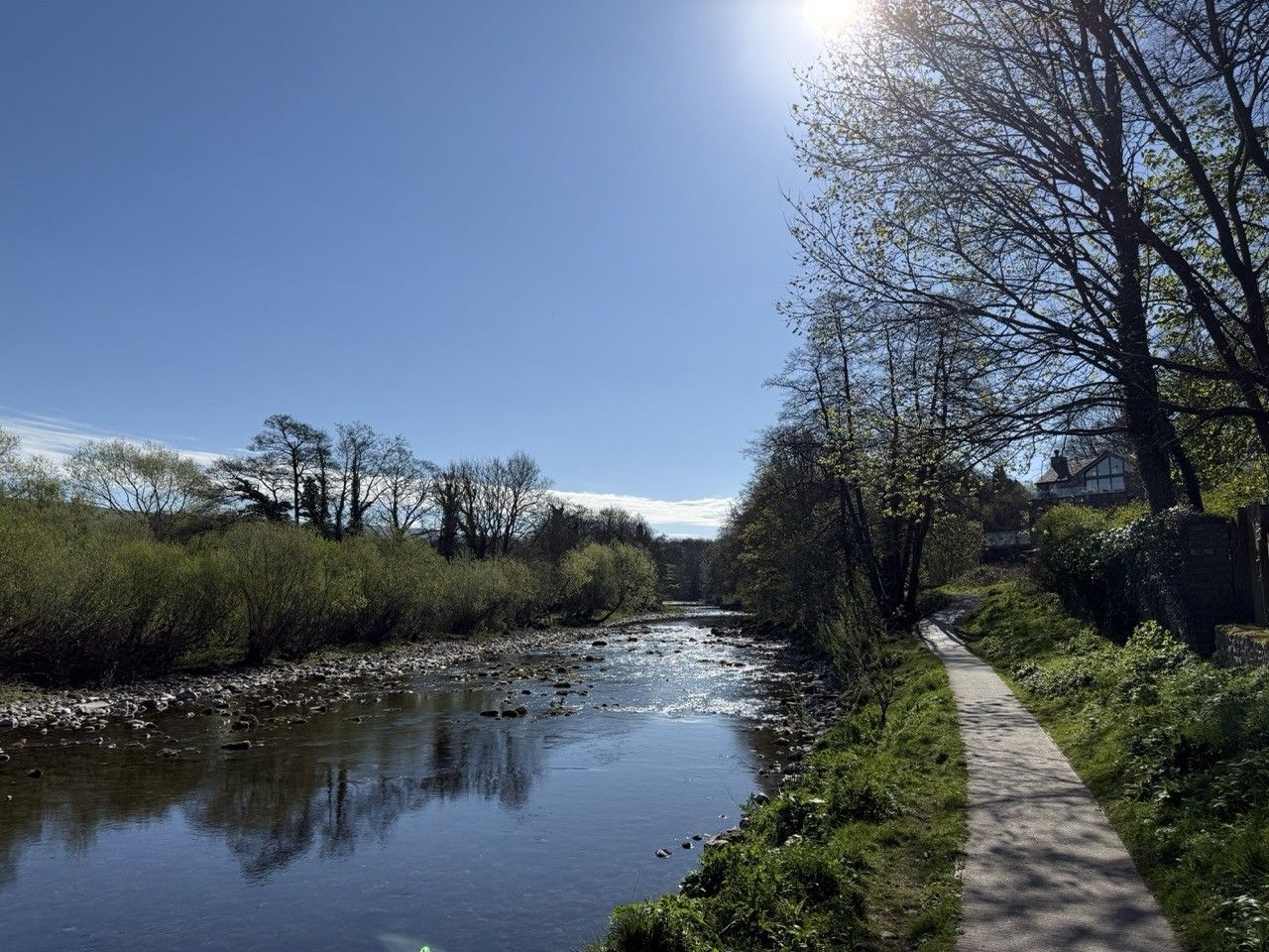 Photograph of a river on a bright sunny day. There are trees either side of the river and a path running along the right-hand side.